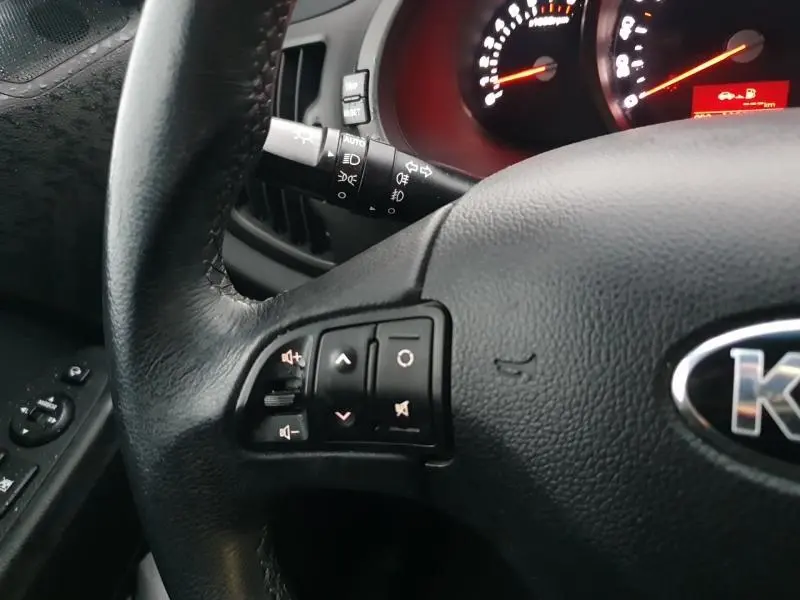 Gros plan sur le volant cuir noir de la KIA SPORTAGE 2013 avec commandes audio et tableau de bord éclairé rouge.