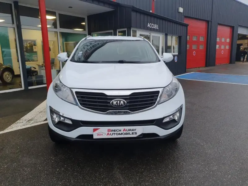 Vue frontale d'un KIA SPORTAGE blanc 2013 avec calandre noire et logo KIA bien visible, stationné devant un bâtiment.