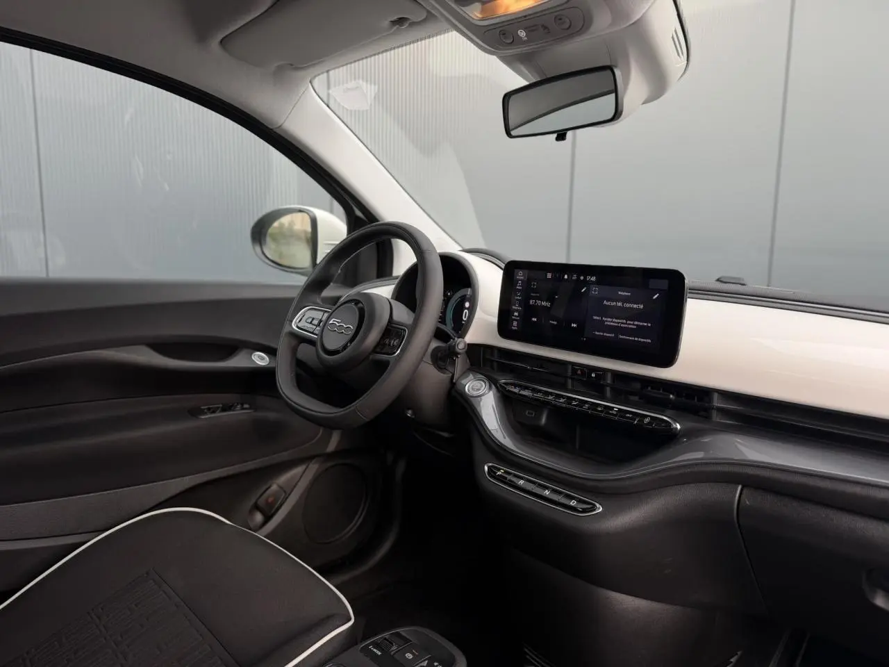Vue intérieure côté conducteur de la Fiat 500 électrique 2024, tableau de bord blanc avec écran tactile et volant noir.