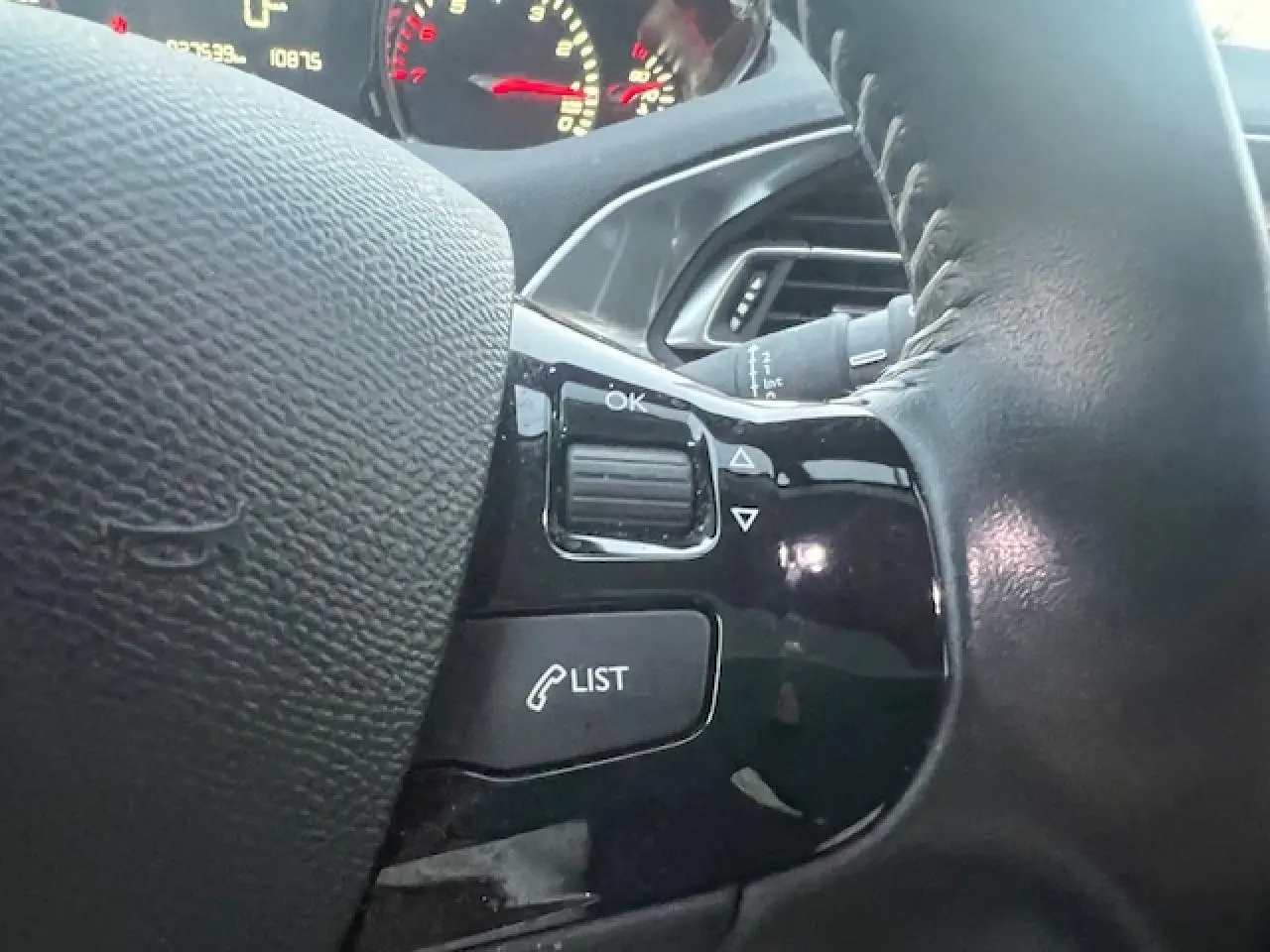 Gros plan sur les commandes au volant noir de la Peugeot 308 blanche, avec tableau de bord partiellement visible en arrière-plan.