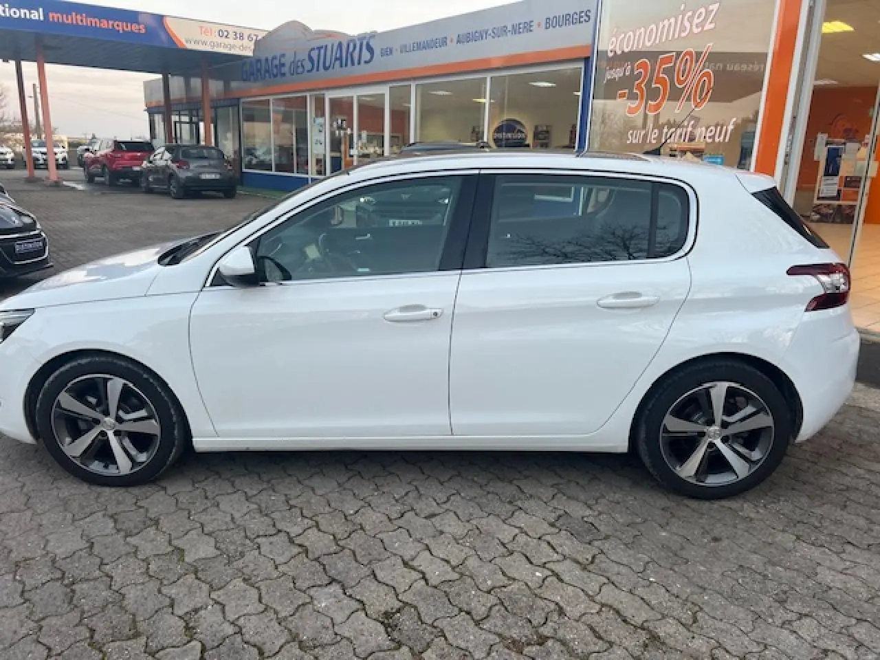 Peugeot 308 blanche vue de profil côté gauche, stationnée devant un garage avec toit panoramique visible.