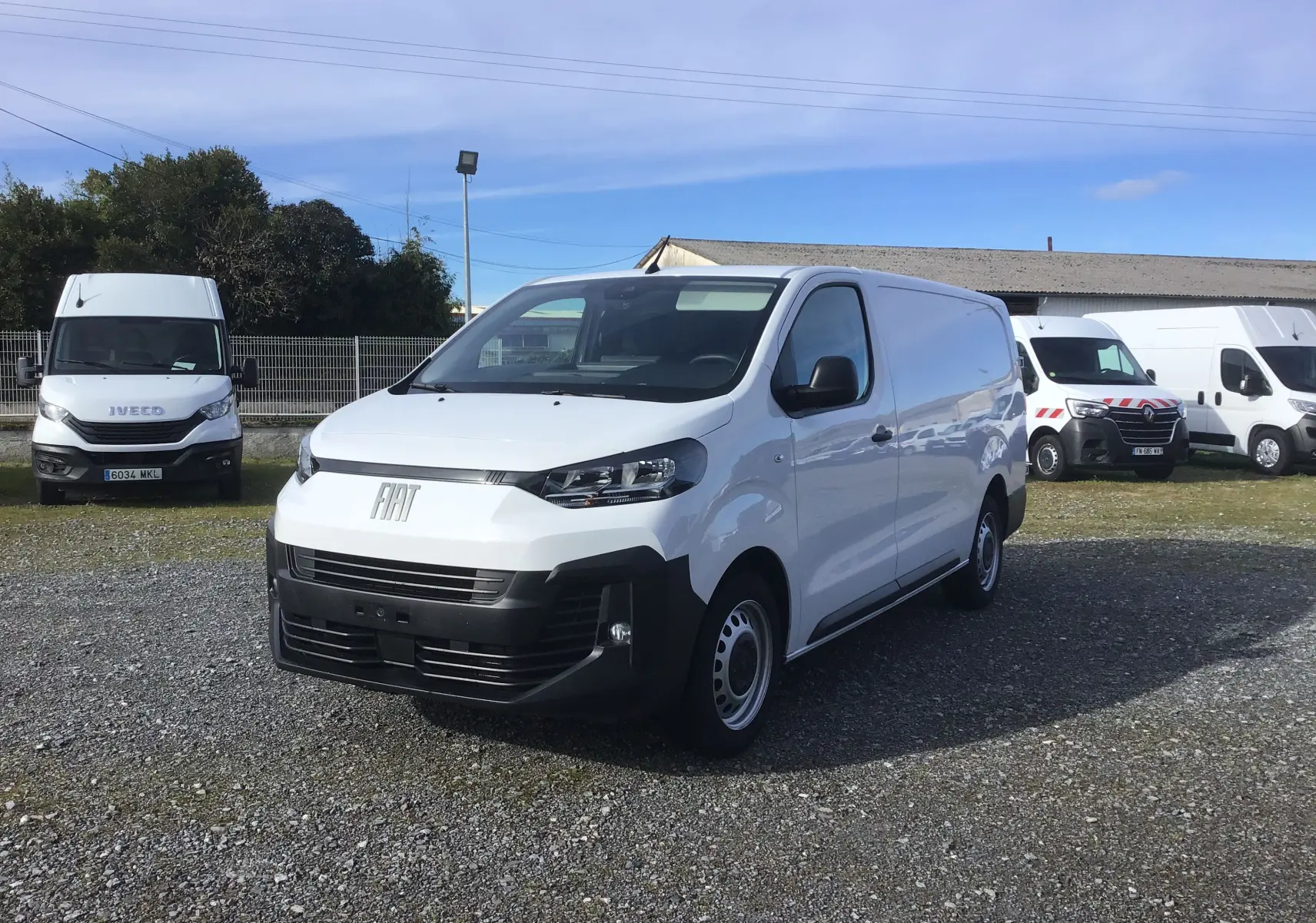 Fiat Scudo Fourgon blanc en 3/4 avant droit, avec pare-chocs noir et calandre moderne, stationné sur un terrain gravillonné.