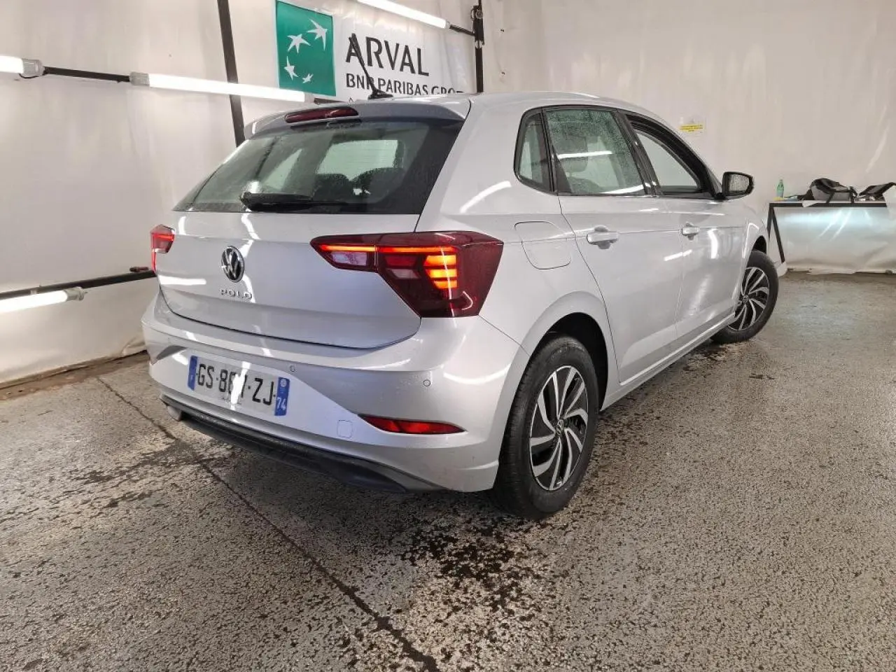 Vue 3/4 arrière droite d'une Volkswagen Polo gris clair avec feux arrière allumés dans un garage.