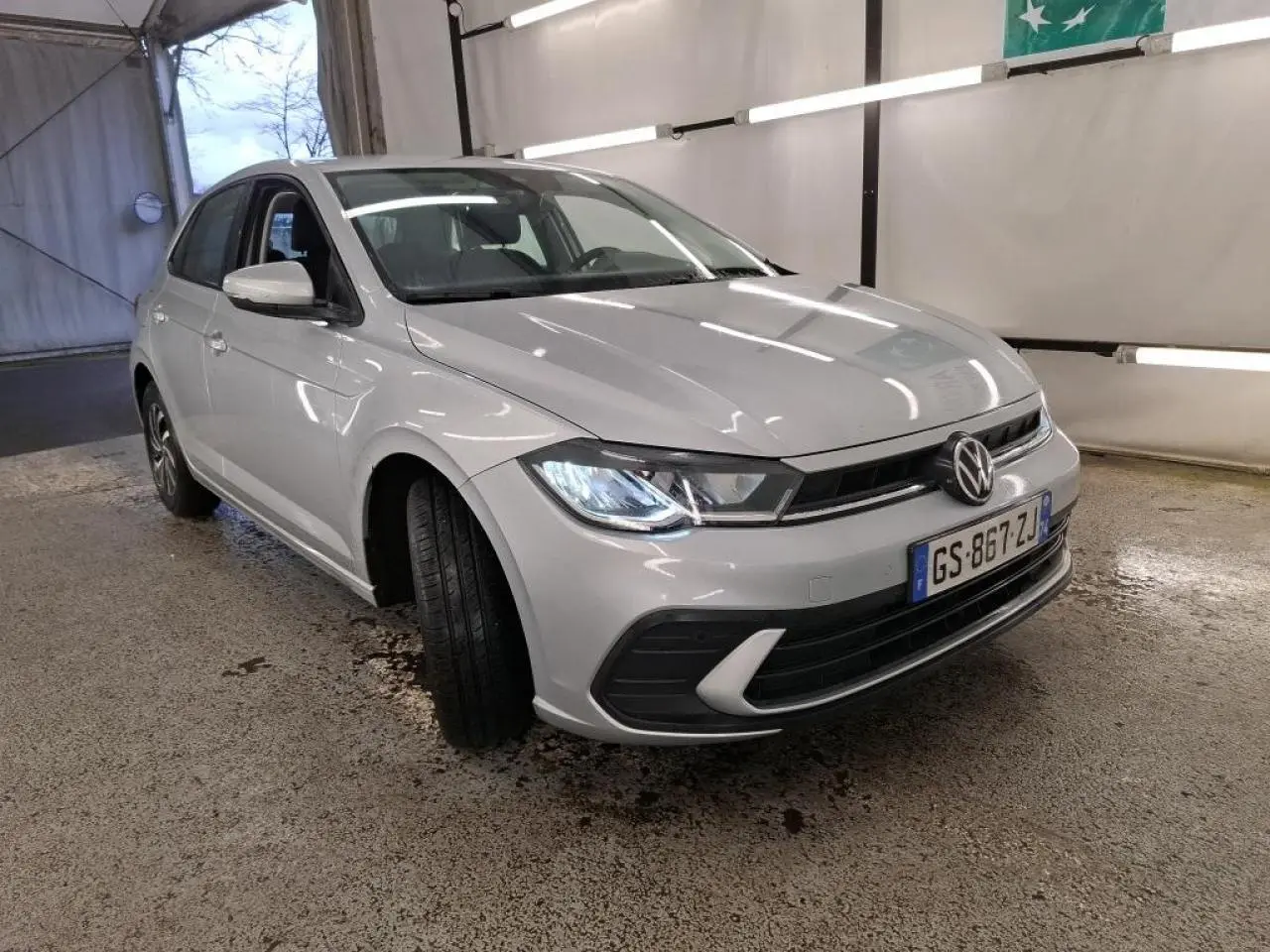 Volkswagen Polo gris clair vue 3/4 avant droit avec phares allumés dans un garage intérieur.