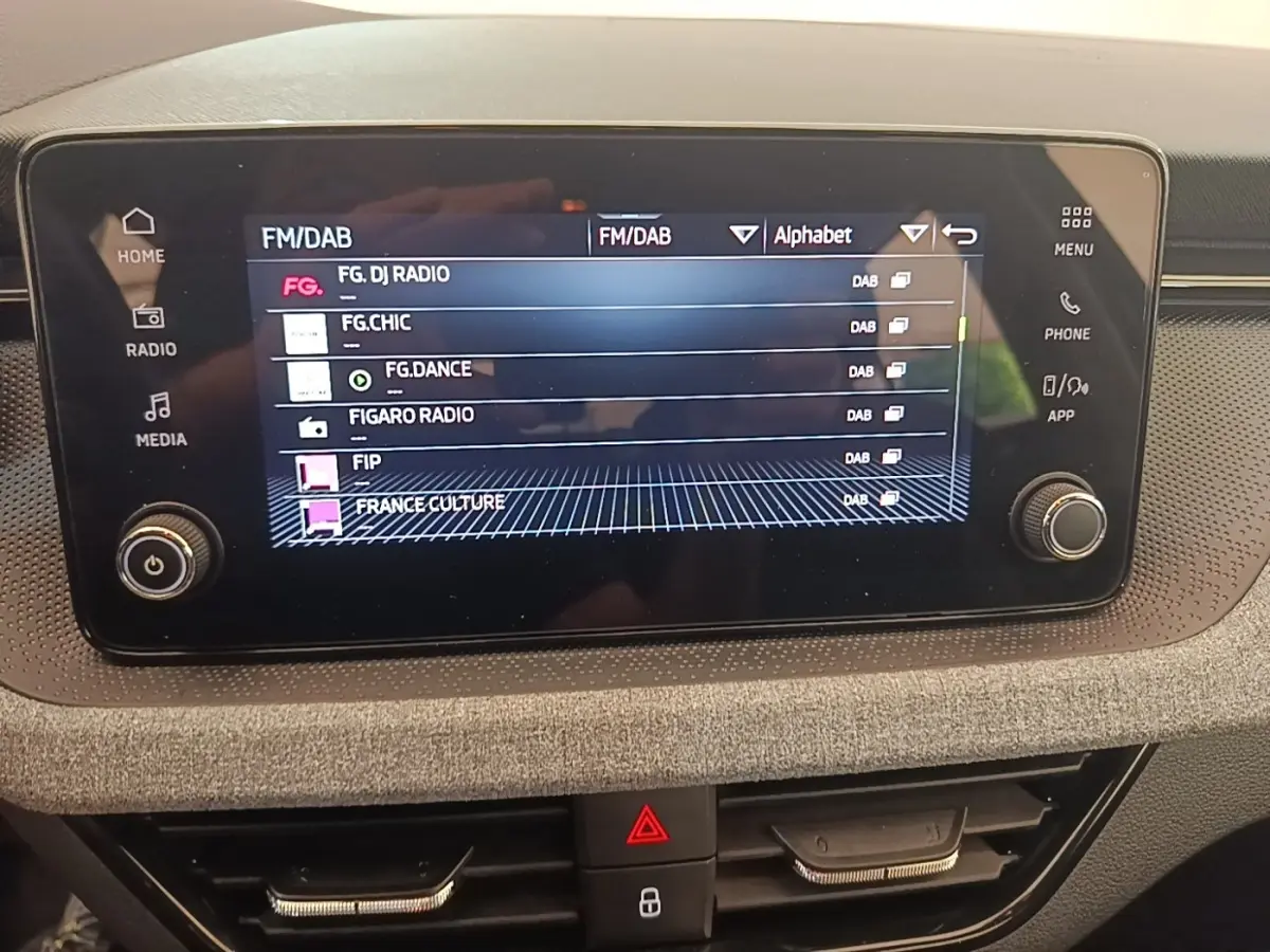 Écran tactile central de la Skoda Scala 2024 affichant les stations radio FM/DAB, entouré de commandes et finition tissu grise.