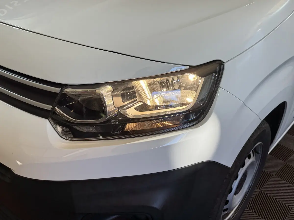 Gros plan sur le phare avant droit allumé d’un Citroën Berlingo blanc avec calandre noire et jante acier.