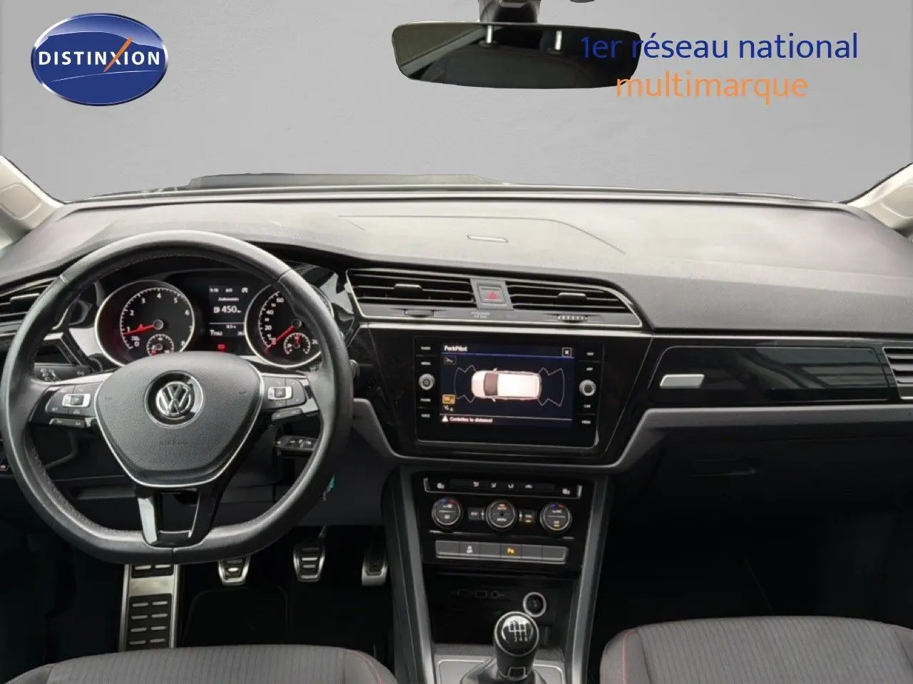 Vue intérieure frontale du tableau de bord et volant noir d’un Volkswagen Touran blanc avec boîte manuelle et écran central tactile.