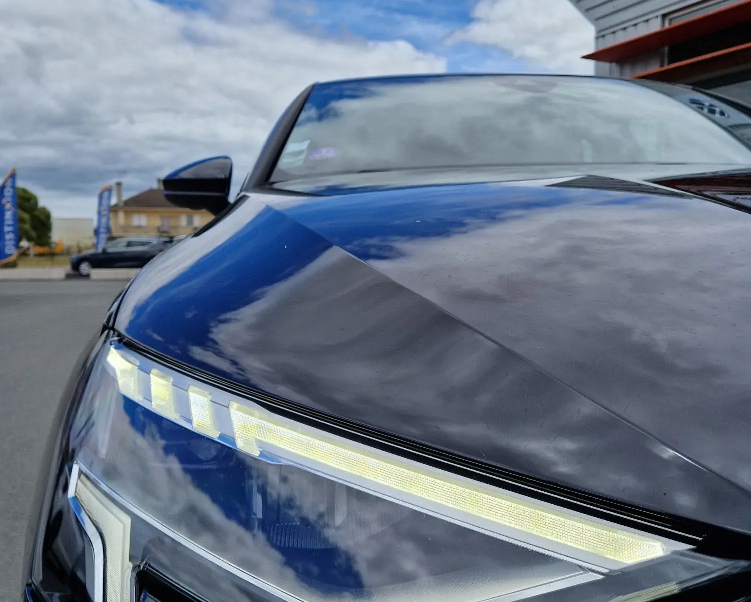 Gros plan sur le phare avant gauche LED d'une Audi A3 Sportback noire, reflétant un ciel nuageux.