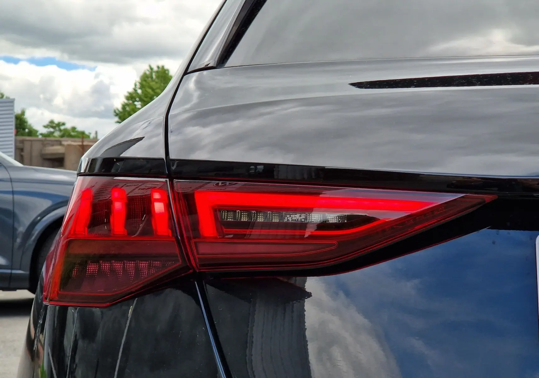 Gros plan sur le feu arrière droit rouge d'une Audi A3 Sportback noire, reflet du ciel nuageux sur la carrosserie.