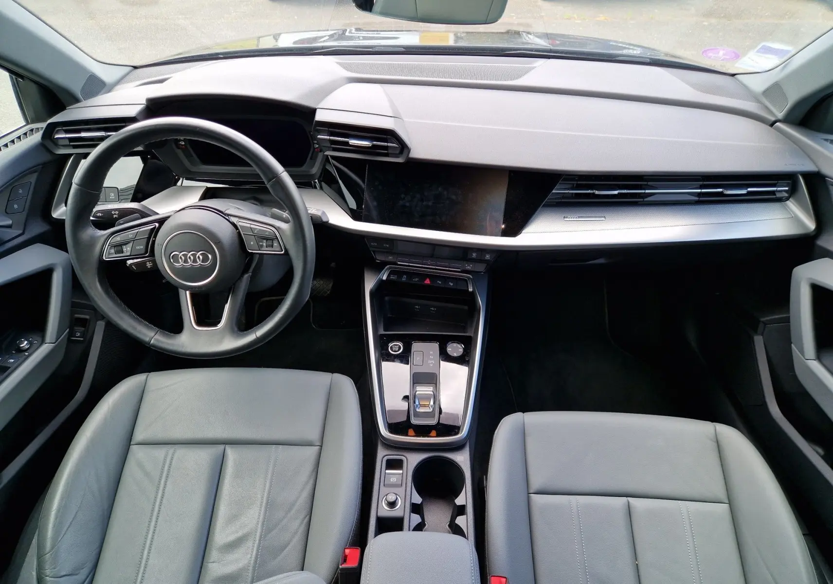 Vue intérieure avant de l'Audi A3 Sportback 40 TFSI-e 2021, tableau de bord moderne avec volant multifonction noir et sièges cuir gris.