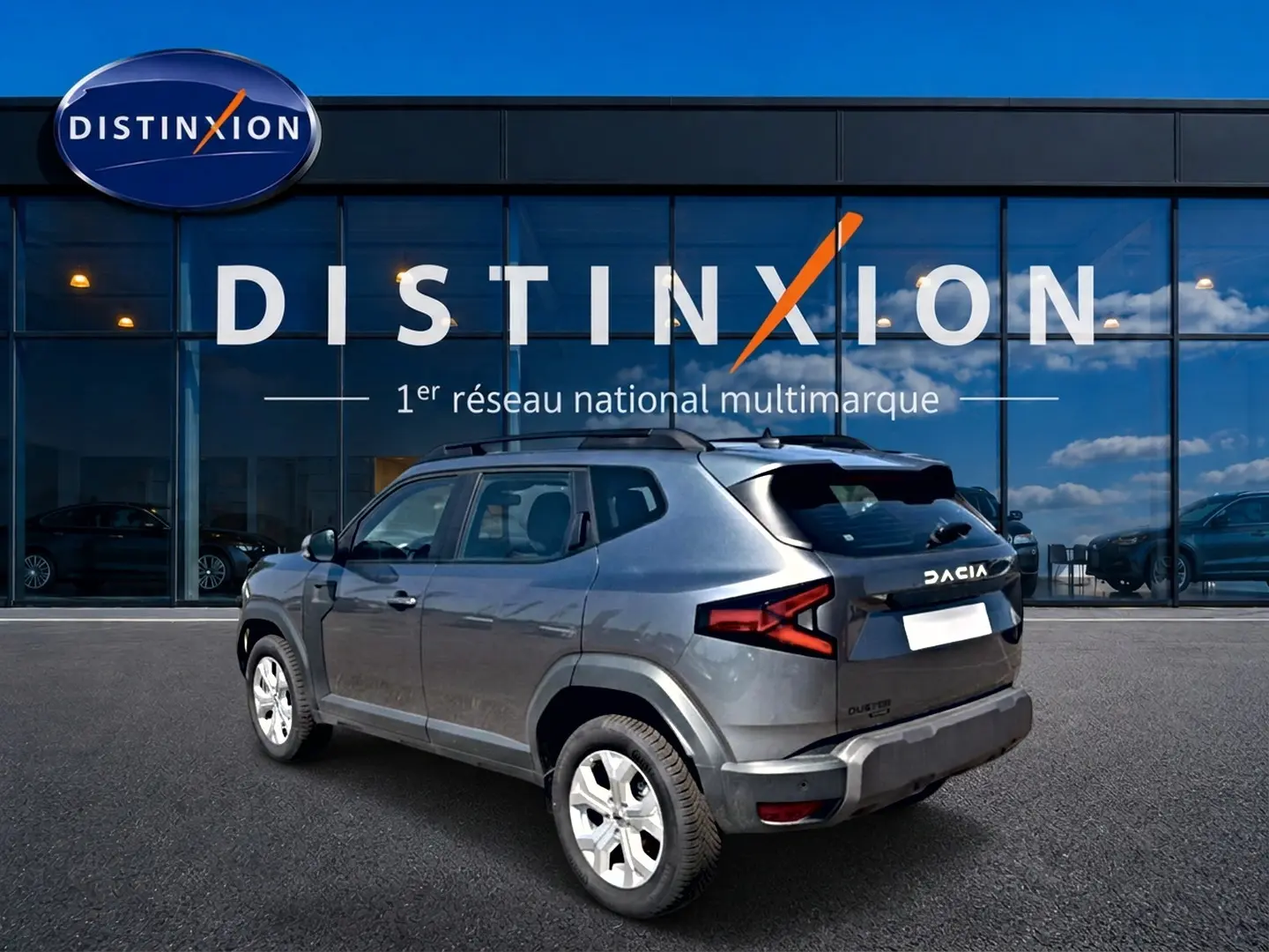 Vue 3/4 arrière droite d'un Dacia Duster gris schiste 2026 avec barres de toit et jantes alu devant un showroom Distinxion.
