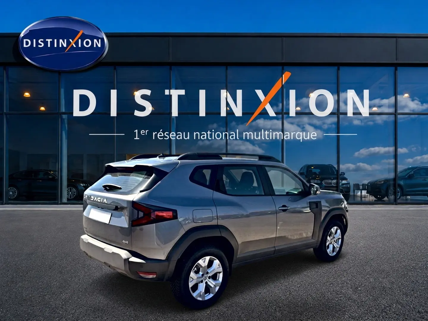 Dacia Duster gris schiste en 3/4 arrière droit, avec barres de toit et jantes alu visibles devant un bâtiment Distinxion.