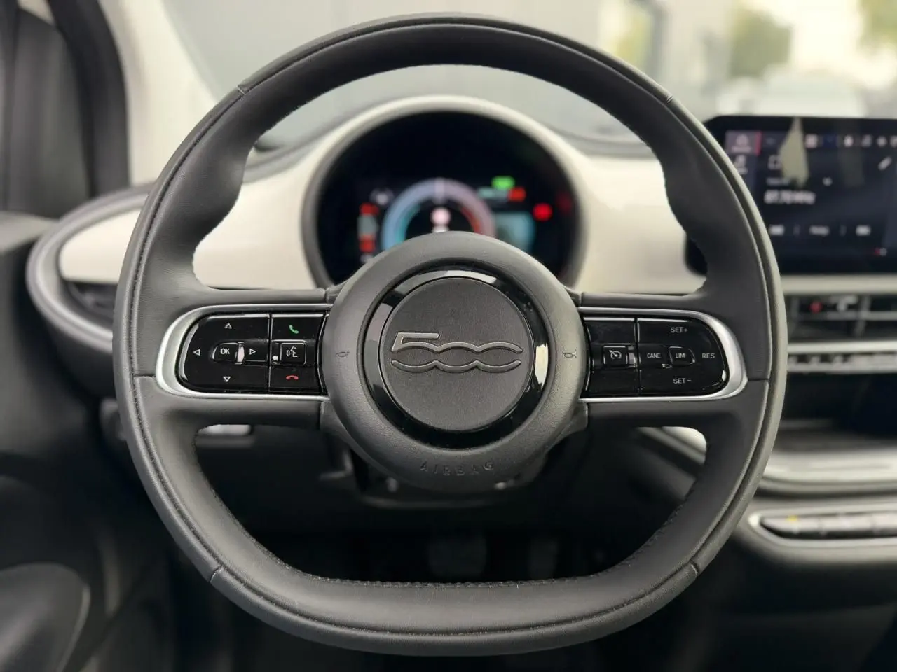 Vue rapprochée du volant noir de la Fiat 500 électrique 2024, avec commandes intégrées et tableau de bord flou en arrière-plan.