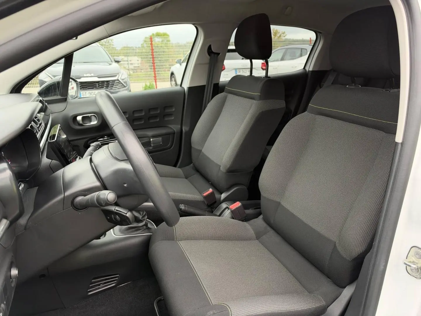 Vue intérieure côté conducteur d'une Citroën C3 blanche, montrant les sièges tissu gris et la console centrale.
