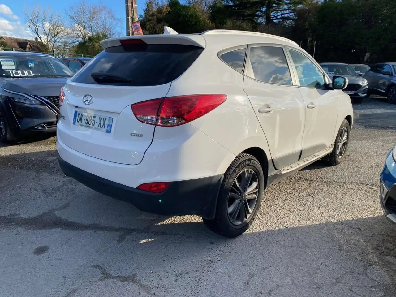 Hyundai ix35 blanc vu en 3/4 arrière droit, avec jantes alliage et vitres teintées sur parking extérieur.