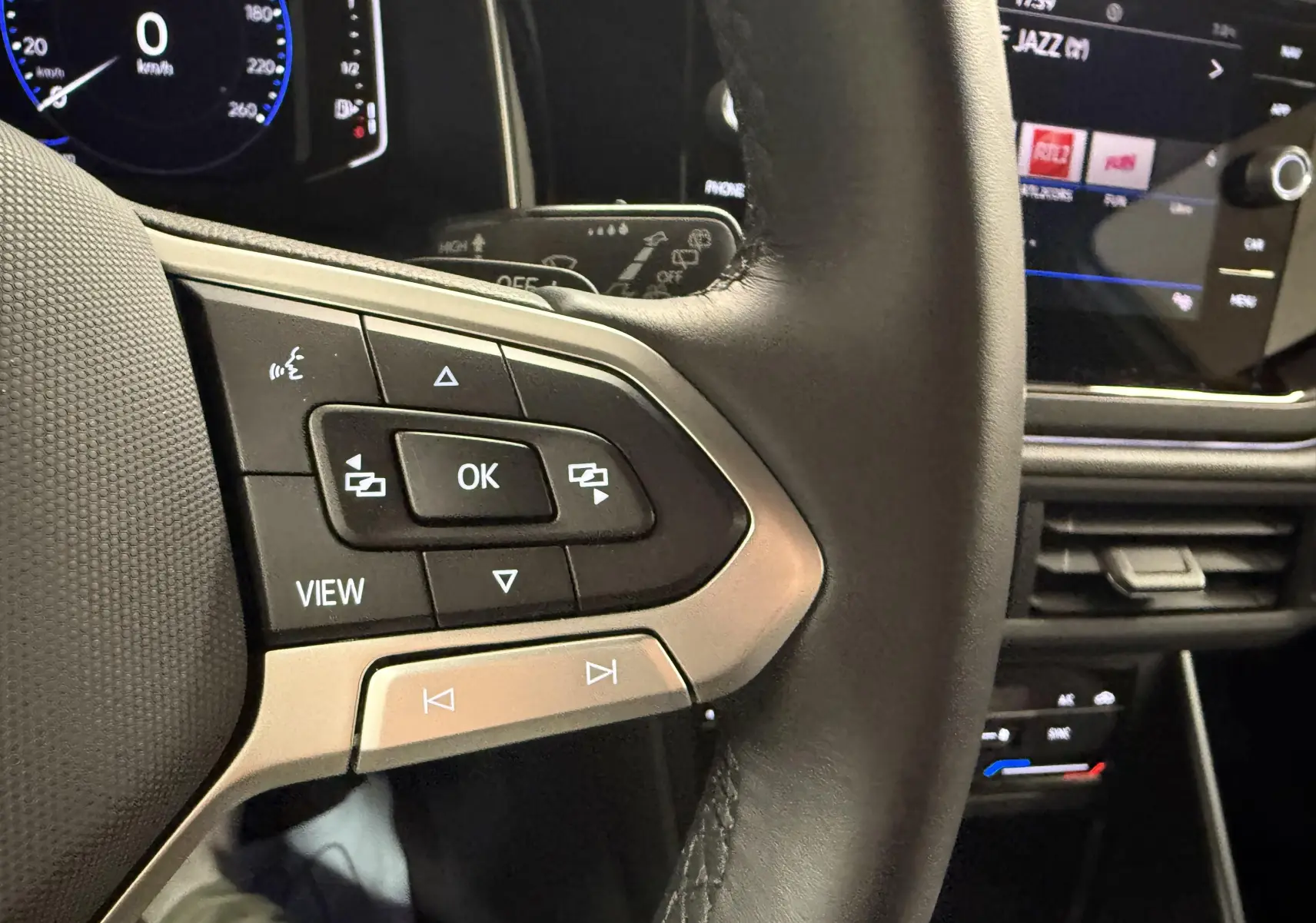 Gros plan sur les commandes gauche du volant en cuir noir du Volkswagen Taigo 2025, avec tableau de bord numérique en arrière-plan.