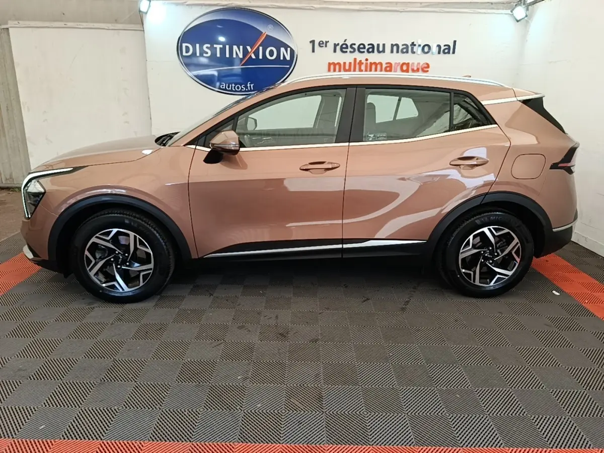 Vue latérale droite d'un KIA SPORTAGE beige 2022 avec jantes alliage et rails de toit noirs en intérieur showroom.