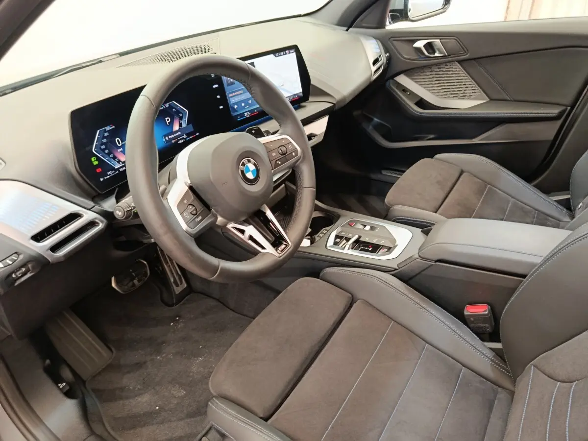 Vue intérieure côté conducteur de la BMW Série 1 120 DKG7 M Sport 2025, montrant volant cuir, écran tactile et sièges gris foncé.