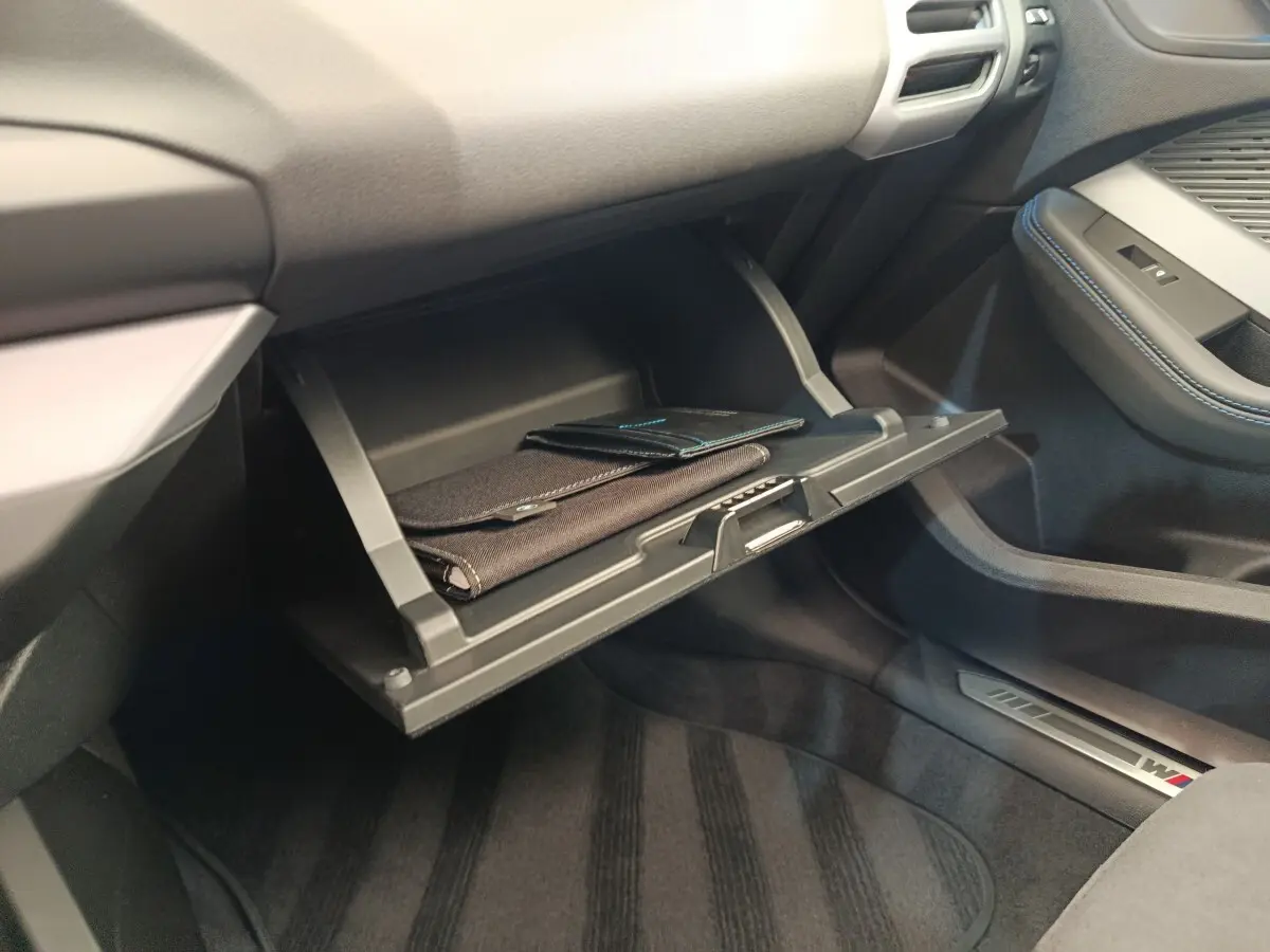 Intérieur BMW Série 1 120 DKG7 M Sport 2025, vue sur boîte à gants ouverte avec documents, sellerie noire et surpiqûres bleues.