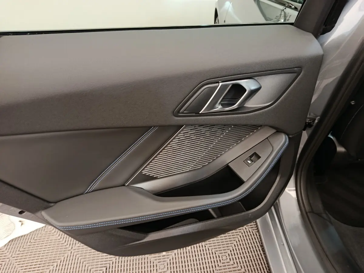 Gros plan sur la porte intérieure côté gauche gris clair de la BMW Série 1 120 DKG7 M Sport avec surpiqûres bleues et poignée métallique.