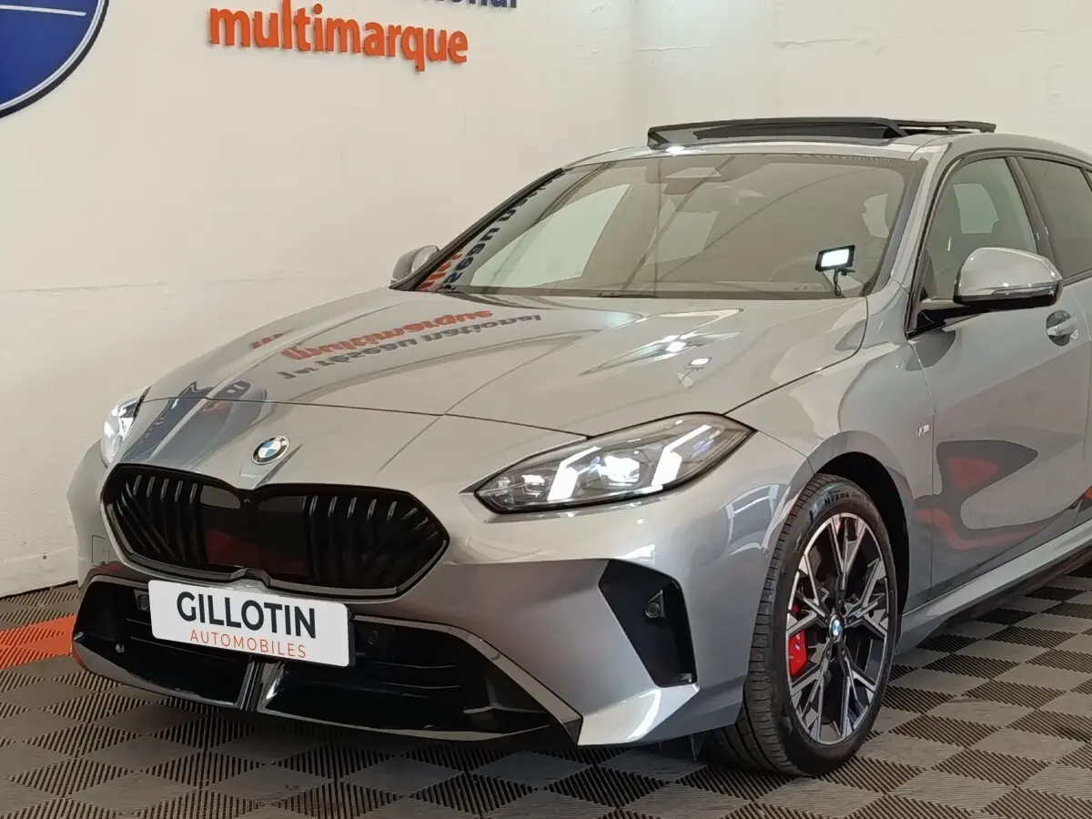 BMW Série 1 120 DKG7 M Sport gris clair vue 3/4 avant avec calandre noire et jantes noires à étriers rouges.