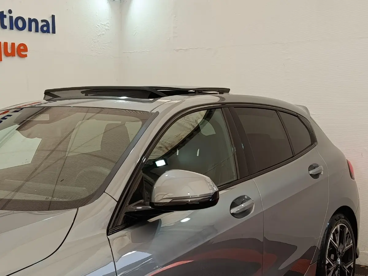 Vue de profil droit avant d'une BMW Série 1 120 DKG7 M Sport gris clair avec toit ouvrant et jantes sport noires.