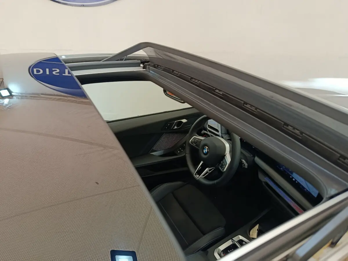 Vue intérieure par le toit ouvrant d'une BMW Série 1 120 DKG7 M Sport gris clair, volant multifonctions et tableau de bord numérique.