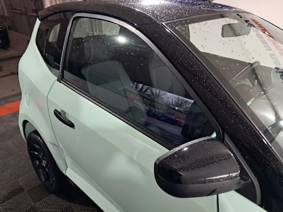 Vue côté conducteur en gros plan d'une Aixam Minauto E chic 2026 verte avec rétroviseur caméra noir et pluie visible.