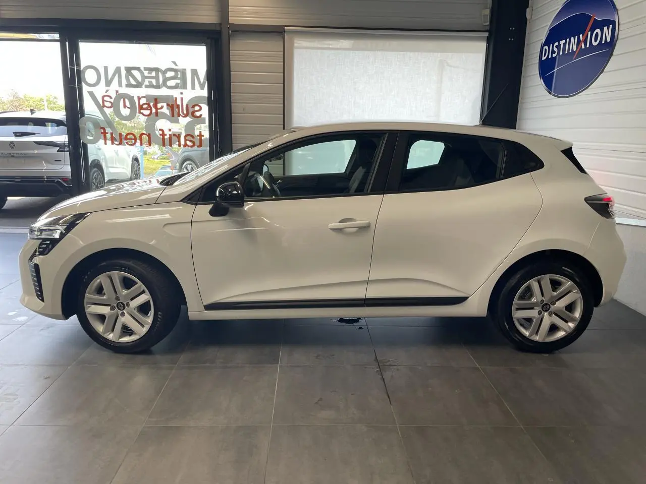 Profil côté gauche d'une Renault Clio blanche 2025 avec jantes alliage et poignées couleur carrosserie dans un showroom.