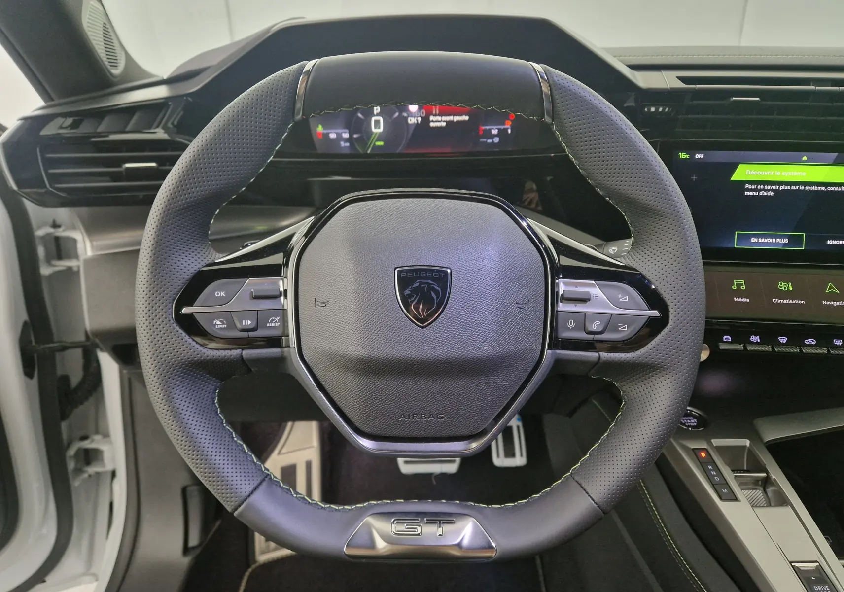 Vue rapprochée du volant cuir perforé noir avec logo Peugeot et badge GT de la 308 blanche 2025, tableau de bord digital visible.