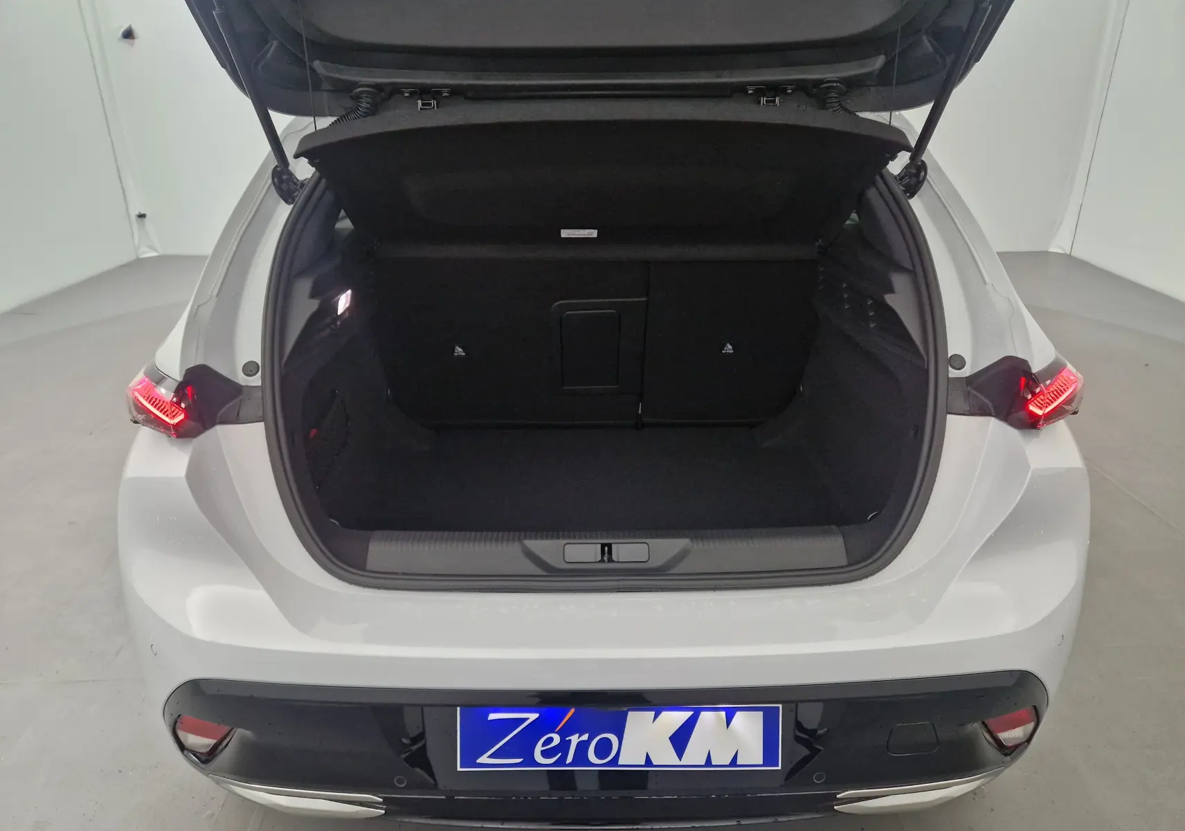 Coffre ouvert vu de l'arrière d'une Peugeot 308 blanche Okenite 2025 avec feux arrière allumés et plaque Zero KM.