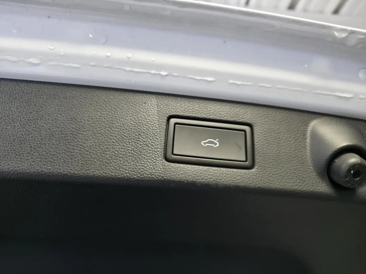 Gros plan sur le bouton de déverrouillage électrique du hayon, entouré de plastique noir, sur Volkswagen Arteon blanc.