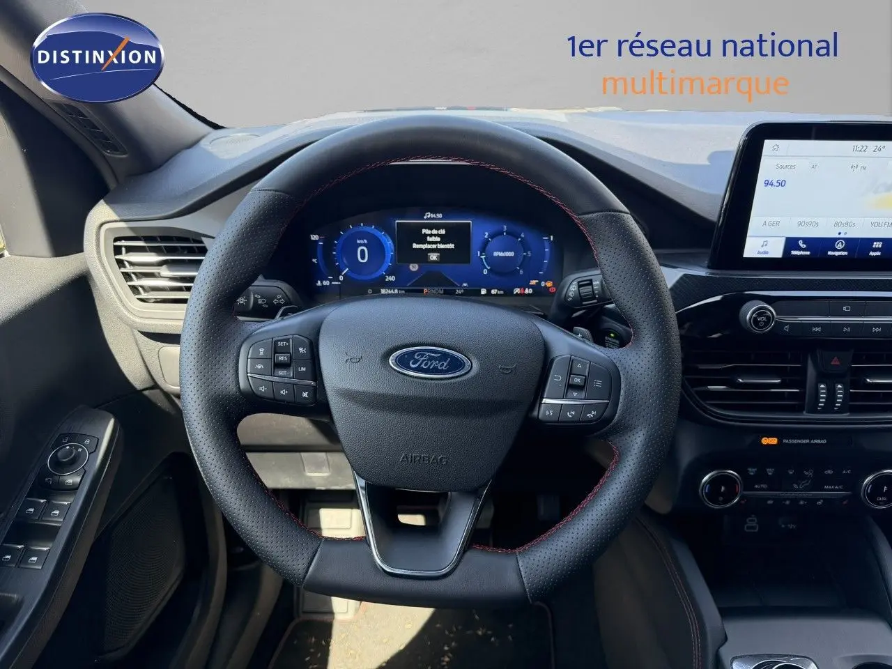Vue intérieure centrée sur le volant cuir noir avec surpiqûres rouges du Ford Kuga 2024, tableau de bord digital et écran tactile.