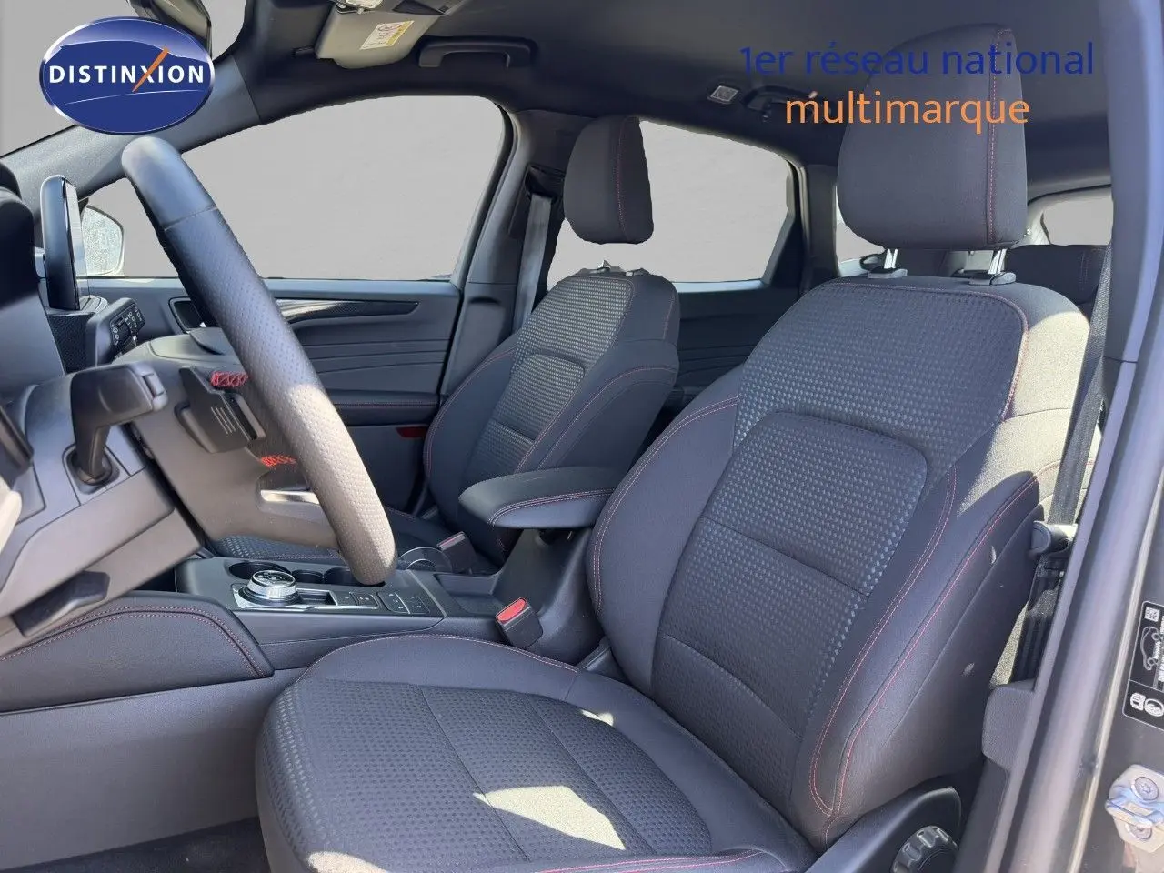 Vue intérieure côté conducteur du Ford Kuga 2024, sièges tissu noir avec surpiqûres rouges et volant multifonction.