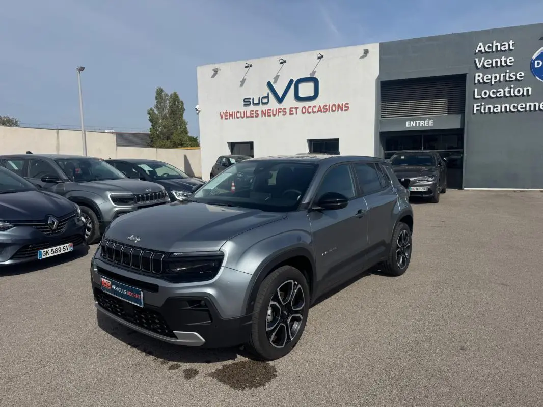 Jeep Avenger gris foncé en 3/4 avant droit, avec calandre noire distinctive et jantes modernes, stationné devant un concessionnaire.