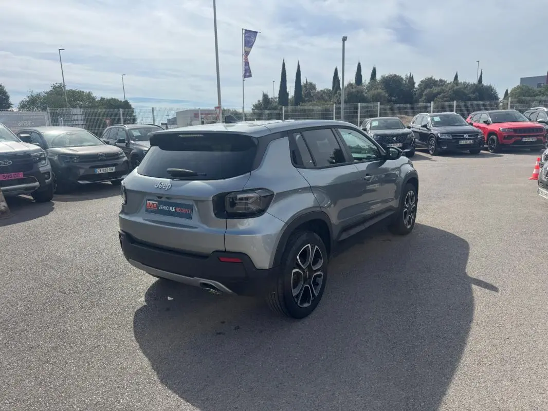 Jeep Avenger gris foncé vu en 3/4 arrière droit sur un parking, avec feux arrière sombres et jantes distinctives.