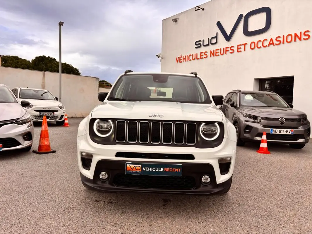 Vue frontale d'un Jeep Renegade blanc 2024 avec calandre noire et phares ronds allumés, stationné devant un concessionnaire.