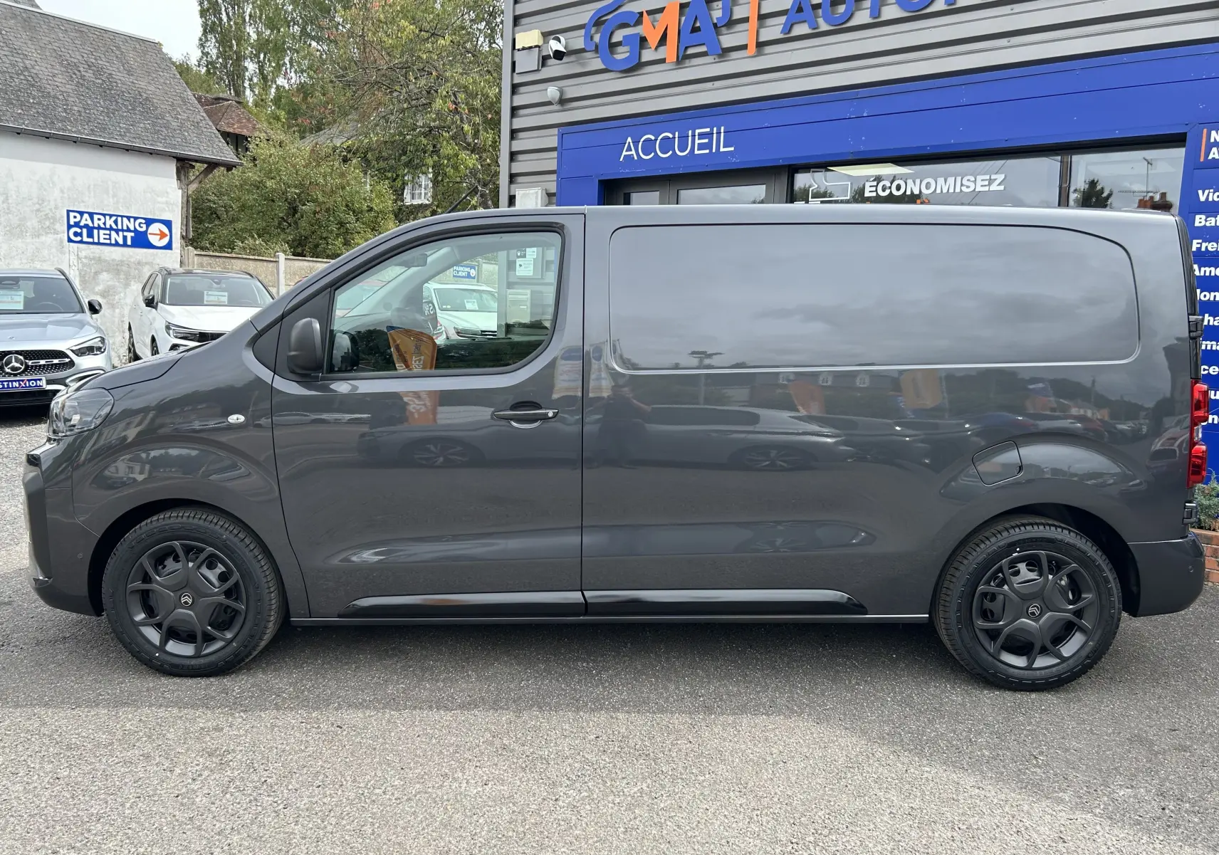 Profil côté gauche d'un utilitaire Citroën Jumpy gris titane 2025 avec jantes noires devant un garage automobile.