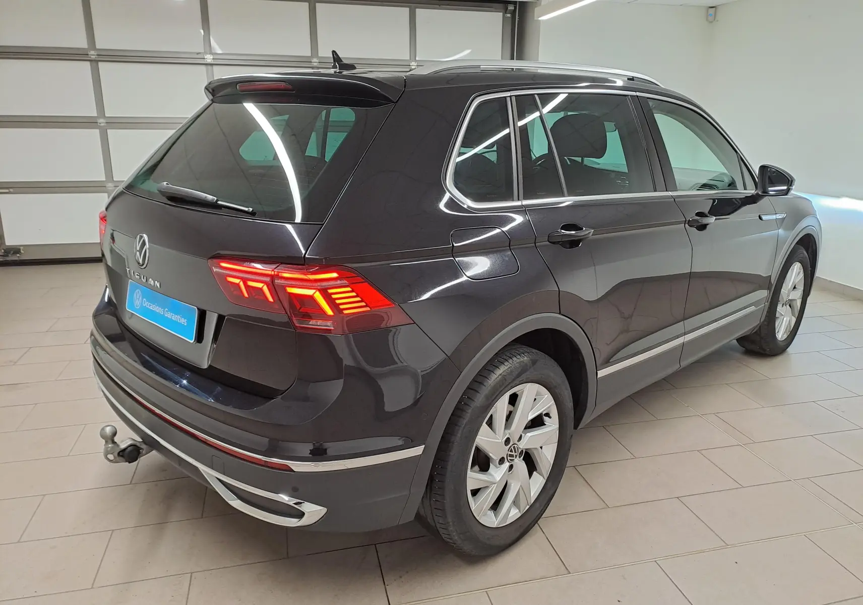 Vue 3/4 arrière droite du Volkswagen Tiguan noir intense 2022 avec feux allumés et crochet d'attelage escamotable visible.