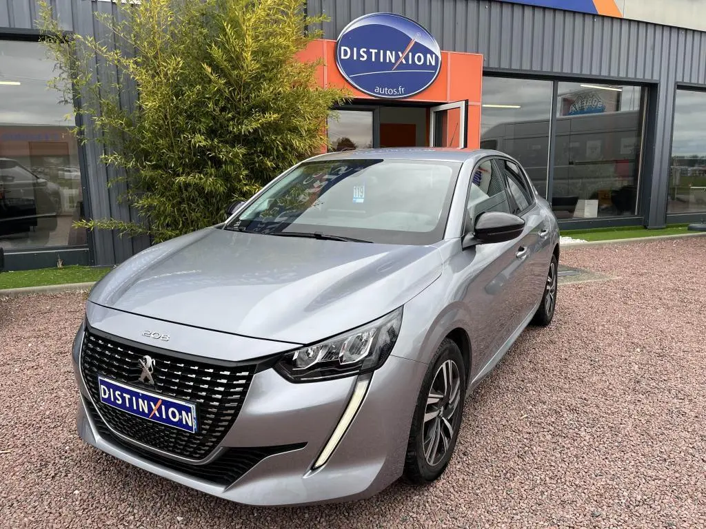 Peugeot 208 gris clair vue 3/4 avant, avec calandre noire et feux LED distinctifs, stationnée devant un bâtiment.