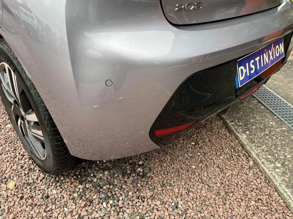 Vue rapprochée du coin arrière gauche d'une Peugeot 208 gris clair avec capteurs de stationnement et logo 208 visible.