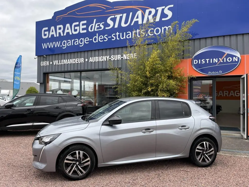 Peugeot 208 gris clair vue de profil côté gauche, stationnée devant un garage avec vitres arrière surteintées.
