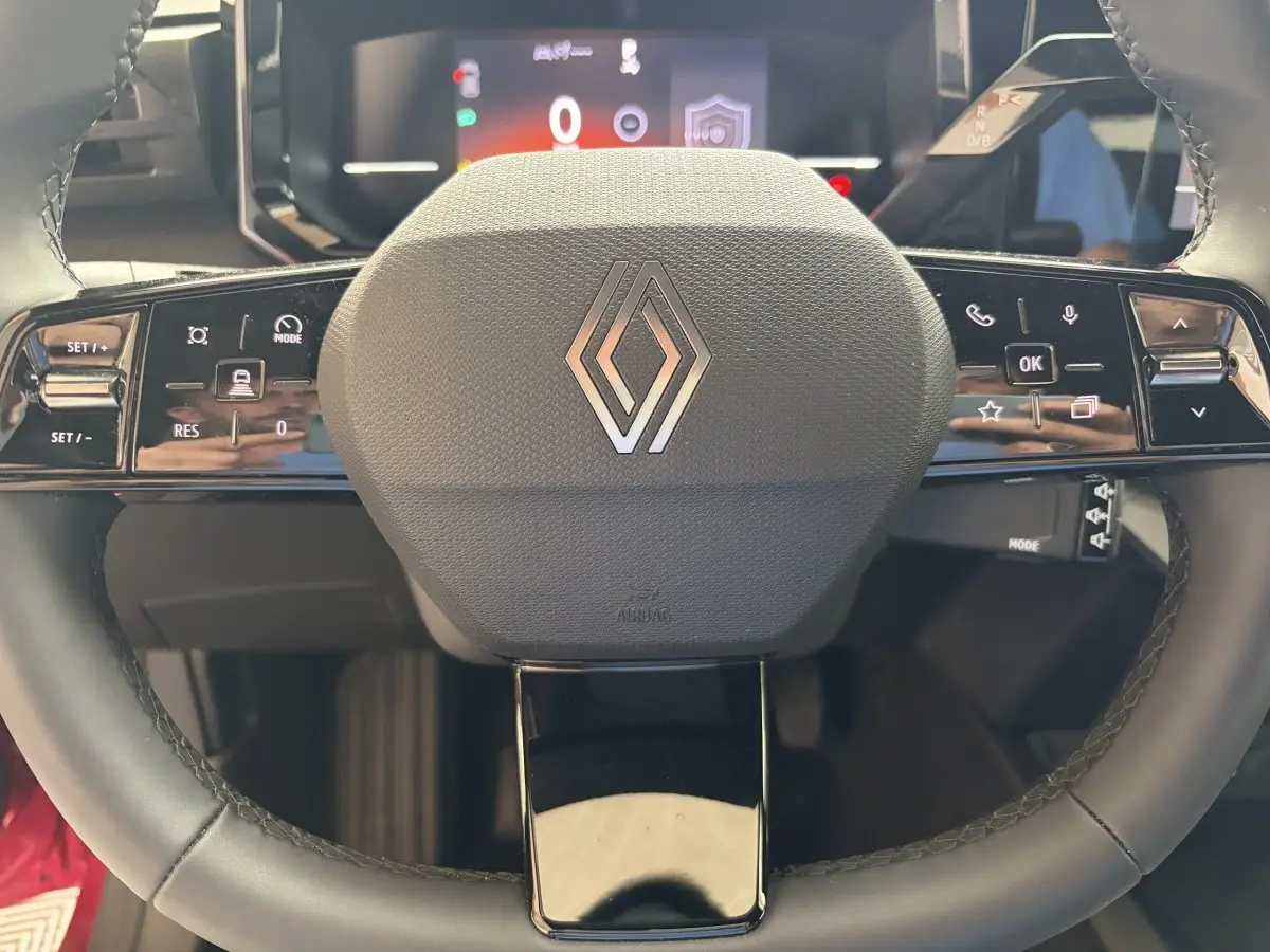 Gros plan sur le volant multifonction noir avec logo Renault, tableau de bord numérique flou en arrière-plan.