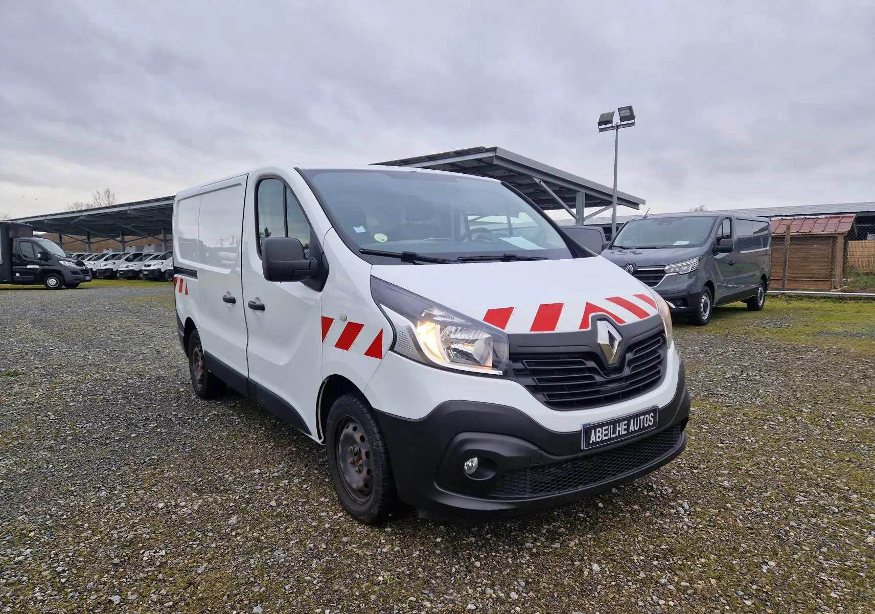 Renault Trafic Fourgon blanc en 3/4 avant droit avec bandes rouges réfléchissantes sur capot et portières.