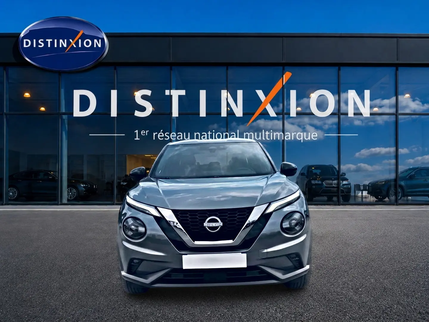 Nissan Juke gris 2025 en vue frontale, avec calandre noire et phares ronds distinctifs, devant showroom Distinxion.