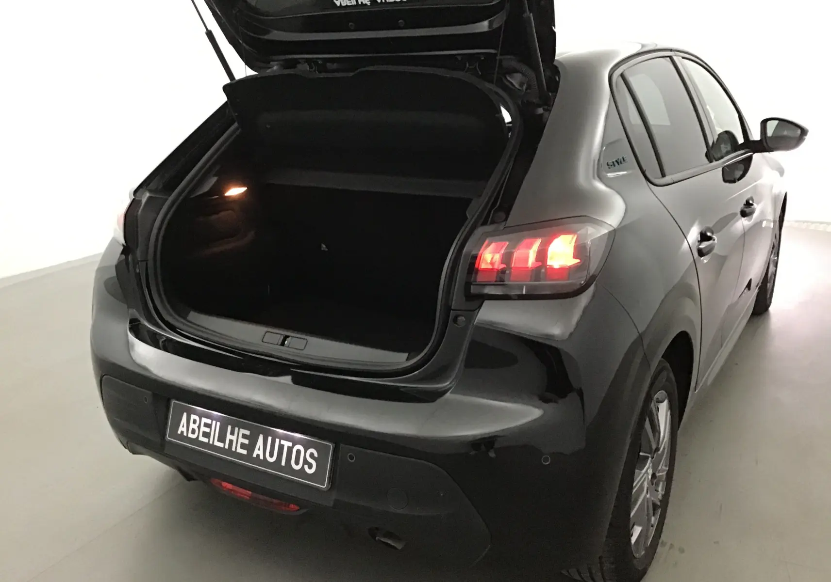 Vue 3/4 arrière droite d'une Peugeot 208 noire 2022 avec coffre ouvert et feux arrière allumés dans un garage.