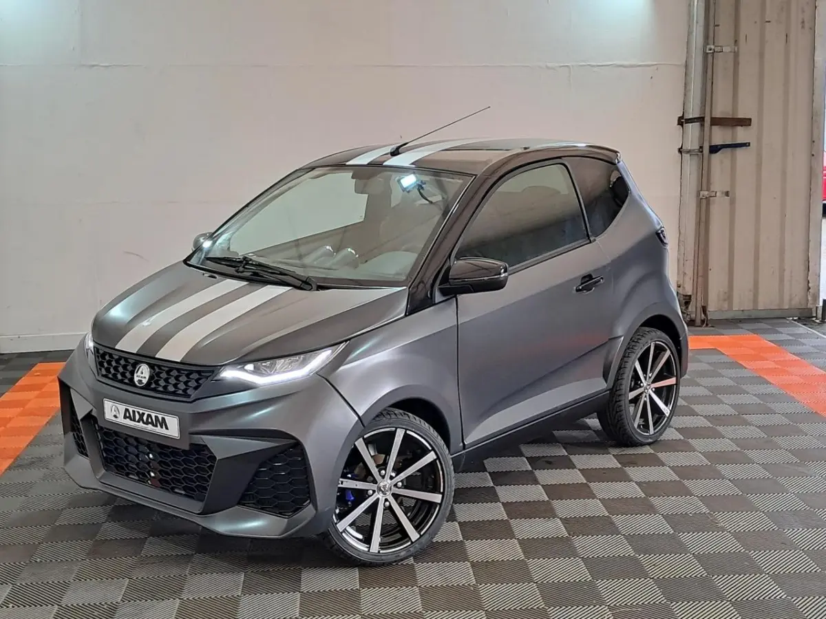 Vue 3/4 avant droite d'une Aixam Ambition gris clair avec bandes blanches, jantes alliage noires diamantées et clignotants à défilement.