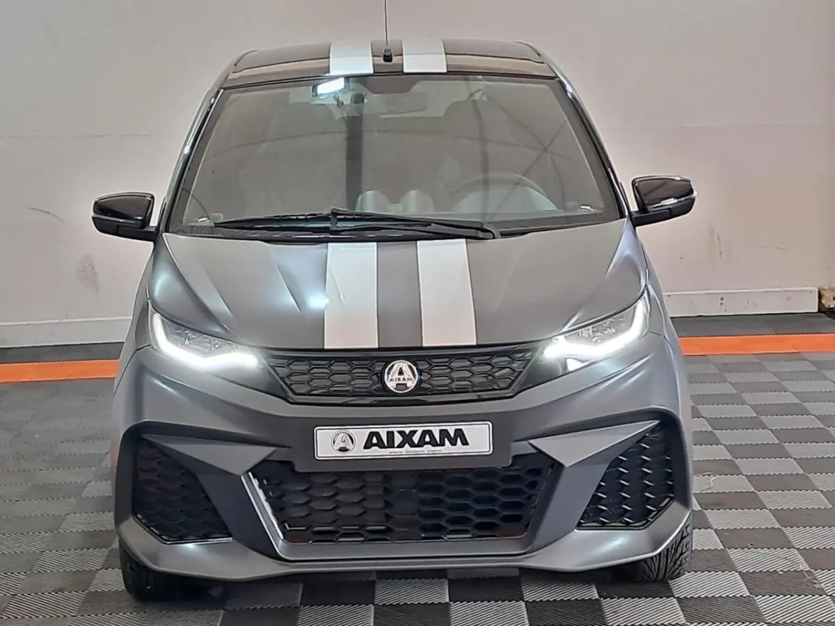 Vue avant d'une Aixam Ambition 2024 gris clair avec bandes blanches et phares LED allumés.