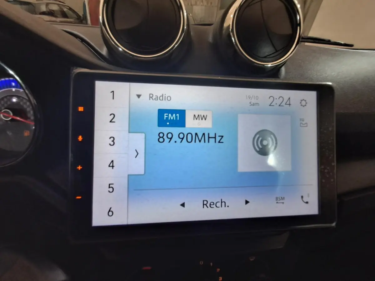 Écran tactile central de l’Aixam Ambition 2024 affichant la radio FM à 89,90 MHz, avec aérateurs ronds au-dessus.