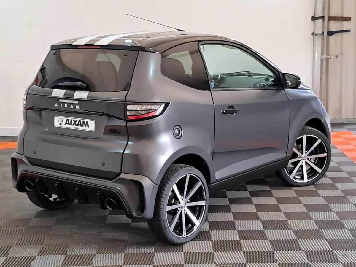 Vue 3/4 arrière droite de l’Aixam Ambition gris clair avec jantes alliage noires diamantées et bandes blanches sur le toit.